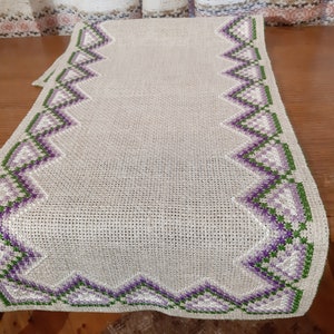 Puede incluir: Un camino de mesa de lino beige con un borde bordado en morado y verde. El borde presenta un patrón repetitivo de triángulos.