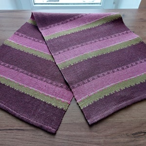 May include: A striped table runner in shades of purple, pink, and green. The runner is woven with a textured pattern and has a fringed edge.