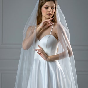 K&ouml;nnte beinhalten: Eine Braut in einem wei&szlig;en Satin-Hochzeitskleid mit einem langen, flie&szlig;enden Schleier. Das Kleid hat einen herzf&ouml;rmigen Ausschnitt und ein tailliertes Oberteil. Der Schleier ist durchsichtig und zart.