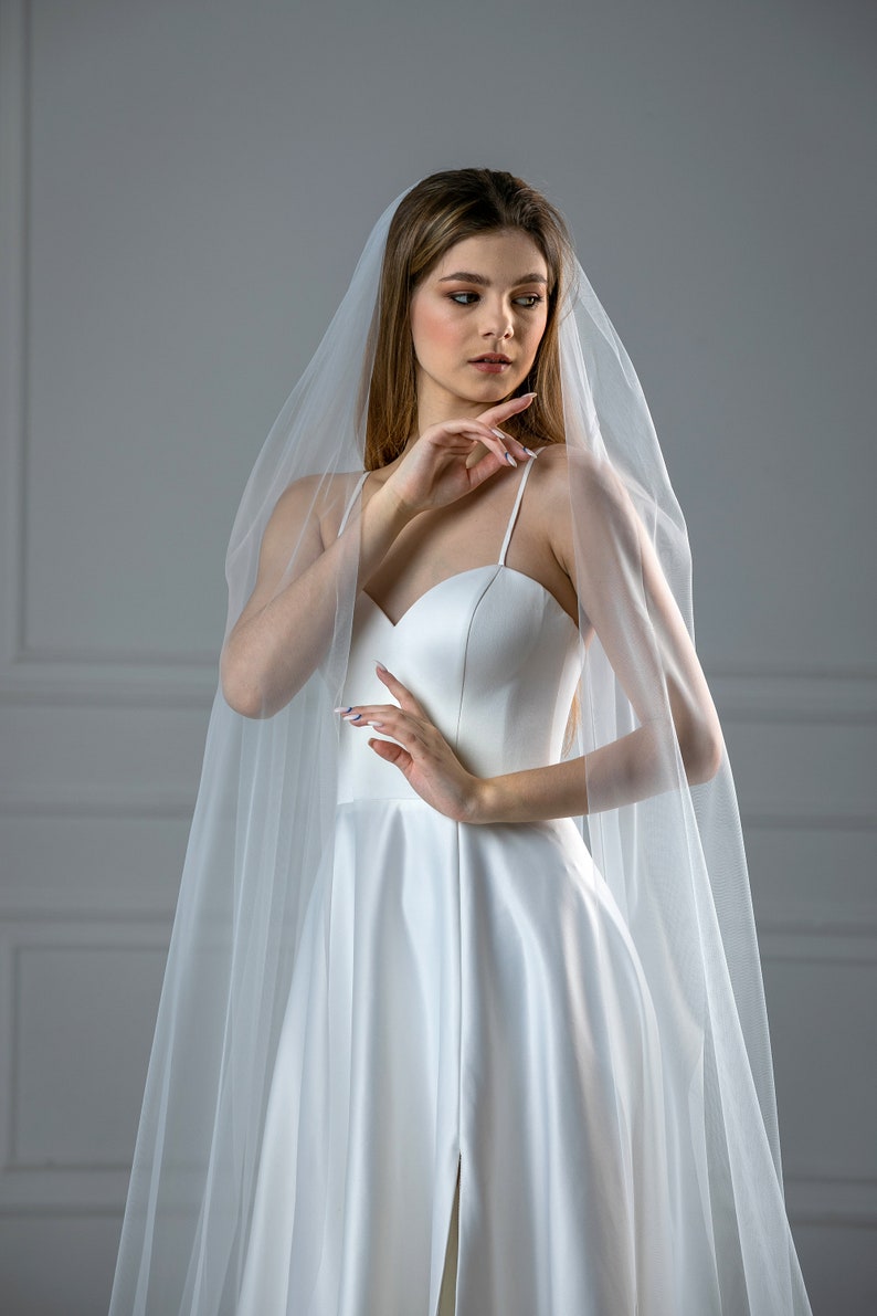 K&ouml;nnte beinhalten: Eine Braut in einem wei&szlig;en Satin-Hochzeitskleid mit einem langen, flie&szlig;enden Schleier. Das Kleid hat einen herzf&ouml;rmigen Ausschnitt und ein tailliertes Oberteil. Der Schleier ist durchsichtig und zart.