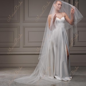 May include: A white bridal gown with a sweetheart neckline and a high slit. The dress is paired with a long, sheer veil. The image is set against a neutral background. The words "Maria Bride" are printed on the image.