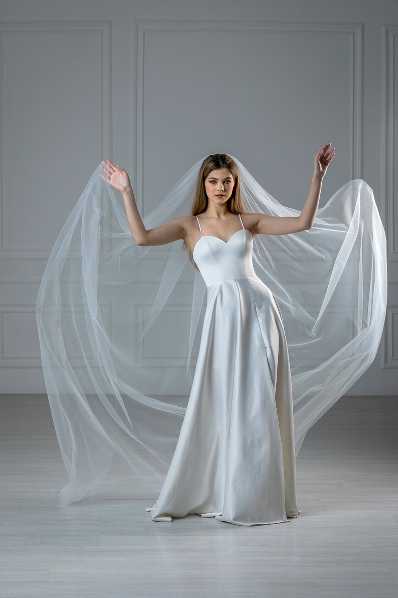 K&ouml;nnte beinhalten: Eine Frau in einem wei&szlig;en Brautkleid mit herzf&ouml;rmigem Ausschnitt und einem langen Schleier. Der Schleier weht hinter ihr, w&auml;hrend sie mit erhobenen Armen posiert.