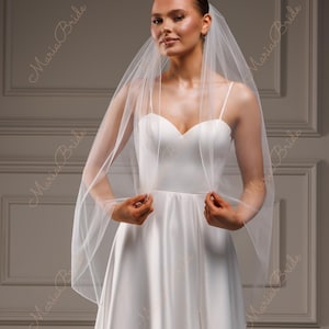 May include: A bride wearing a white satin wedding dress and a sheer veil. The dress has a sweetheart neckline and spaghetti straps. The veil is held up by the bride's hands. The words "Maria Bride" are printed on the image.