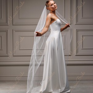 May include: A white bridal gown with a flowing skirt and a simple bodice. The bride is wearing a long, sheer veil. The background is a neutral wall with decorative panels. The image has the text "Maria Bride" in the corners.
