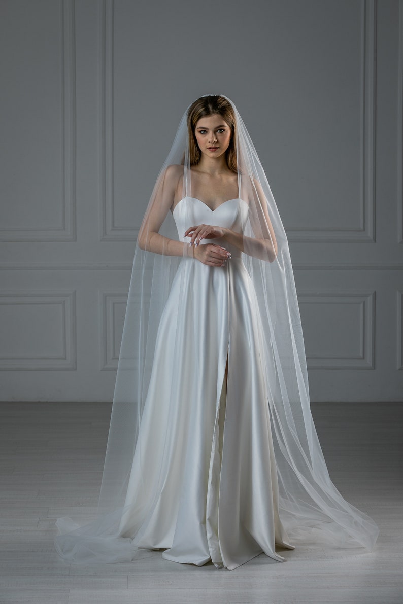 K&ouml;nnte beinhalten: Eine Braut in einem wei&szlig;en Brautkleid mit langem Schleier. Das Kleid hat einen herzf&ouml;rmigen Ausschnitt und einen Schlitz vorne.