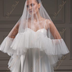 May include: A white wedding dress with a layered tulle bodice and sleeves. The dress features a sweetheart neckline and a long, flowing skirt. A sheer veil drapes over the head and shoulders, creating a romantic and elegant look.