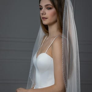 May include: A bride wearing a white wedding dress with a long, sheer veil with a beaded edge. The veil is draped over her head and shoulders.