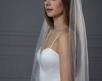 May include: A bride wearing a white wedding dress with a long, sheer veil with a beaded edge. The veil is draped over her head and shoulders.
