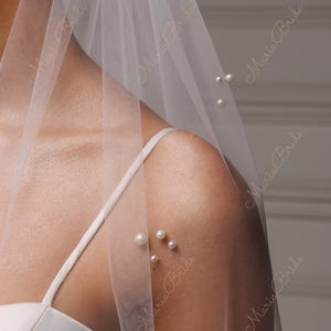 May include: A sheer white bridal veil adorned with scattered faux pearls. The veil is draped over a shoulder, revealing a white spaghetti strap dress. The words "Maria Bride" are printed on the veil.
