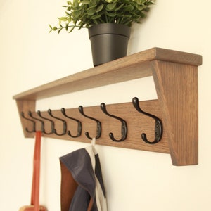 Puede incluir: Un perchero de pared de madera con un estante y seis ganchos de metal negro. Una planta verde en una maceta negra se encuentra en el estante. Una correa de cuero marrón y una chaqueta azul cuelgan de los ganchos.