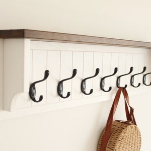 May include: White wooden coat rack with a brown top shelf and eight black metal hooks. A brown leather bag with a woven straw circle is hanging on one of the hooks.