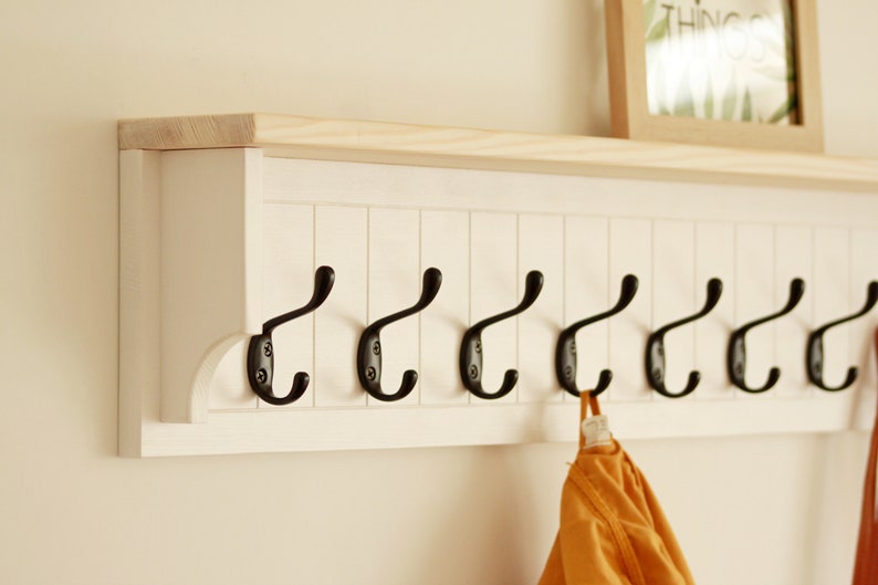 May include: White wooden coat rack with a shelf on top and eight black metal hooks. A yellow garment is hanging on one of the hooks.