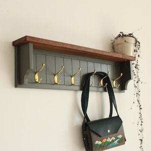 May include: A gray wooden wall shelf with a brown top shelf and five gold hooks. A black leather bag hangs from one of the hooks.