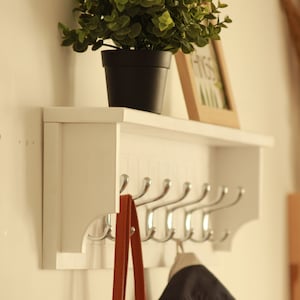 May include: White wall mounted coat rack with a shelf. The rack has six hooks and a small potted plant on the shelf. A brown bag and a blue jacket are hanging on the hooks.