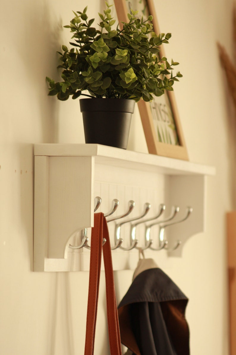 May include: White wall mounted coat rack with a shelf. The rack has six hooks and a small potted plant on the shelf. A brown bag and a blue jacket are hanging on the hooks.