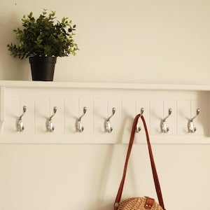 Puede incluir: Perchero de madera blanco con un estante y seis ganchos. Una correa de cuero marrón con una bolsa tejida cuelga de uno de los ganchos.