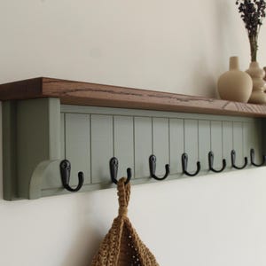 May include: A wall-mounted coat rack featuring a sage green vertical paneled backboard with eight black metal hooks. The unit is topped with a natural dark brown wooden shelf. A tan crocheted bag hangs from one of the hooks below the shelf.