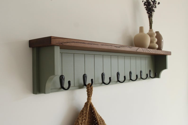 May include: A wall-mounted coat rack featuring a sage green vertical paneled backboard with eight black metal hooks. The unit is topped with a natural dark brown wooden shelf. A tan crocheted bag hangs from one of the hooks below the shelf.