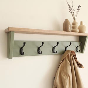 May include: A wall-mounted coat rack featuring a natural wood top shelf and a sage green backboard with five black metal hooks. A tan trench coat hangs from one hook, and three ceramic vases sit on the shelf above the rack.