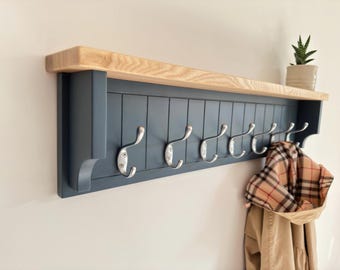 May include: A wall-mounted coat rack with a natural wood shelf and a dark blue base. The rack features seven silver hooks for hanging coats and other items. A plaid scarf and a tan coat hang from the hooks. A small potted plant sits on the shelf.