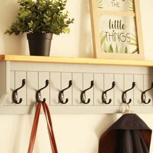 May include: A gray wooden coat rack with a shelf and eight black metal hooks. The shelf has a natural wood finish. A brown leather bag hangs on one of the hooks and a black jacket hangs on another hook. The text "the little things" is visible on a framed picture above the rack.