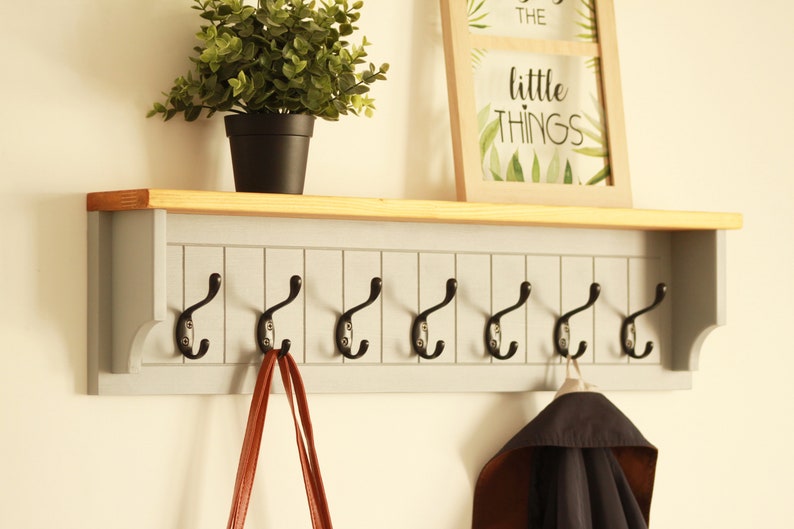 May include: A gray wooden coat rack with a shelf and eight black metal hooks. The shelf has a natural wood finish. A brown leather bag hangs on one of the hooks and a black jacket hangs on another hook. The text "the little things" is visible on a framed picture above the rack.