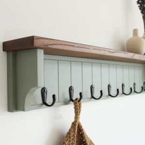 May include: A wall-mounted coat rack featuring a natural wood top shelf and a sage green vertical paneled base. The unit includes eight black metal hooks attached to the front panel, providing storage for hanging items in a home entryway or hallway.