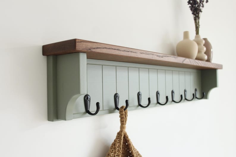May include: A wall-mounted coat rack featuring a natural wood top shelf and a sage green vertical paneled base. The unit includes eight black metal hooks attached to the front panel, providing storage for hanging items in a home entryway or hallway.