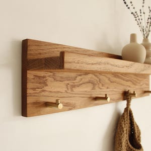 Puede incluir: Estante de pared de madera con acabado natural, con un estante superior para decoración y ganchos de latón debajo. El estante contiene pequeños jarrones y una ramita de flores secas. Un artículo de punto cuelga de uno de los ganchos.