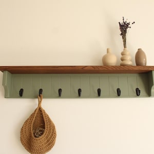 May include: A wall-mounted wooden shelf with a dark brown top surface and a sage green vertical paneled backboard featuring seven black metal hooks. A tan woven hanging basket is suspended from one hook, and three ceramic vases sit on the shelf.