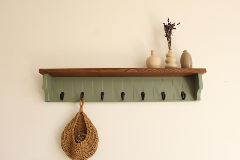 May include: A wall-mounted wooden shelf with a dark brown top surface and a sage green vertical paneled backboard featuring seven black metal hooks. A tan woven hanging basket is suspended from one hook, and three ceramic vases sit on the shelf.