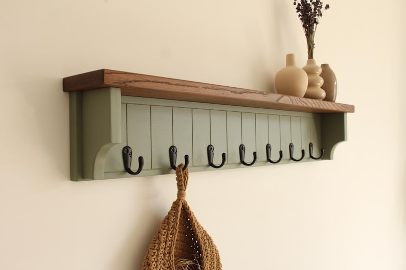 May include: A wall-mounted shelf featuring a natural wood top and a sage green vertical paneled base. The unit includes eight black metal hooks attached to the bottom edge. A woven tan bag hangs from one hook, and three vases sit on the top shelf.