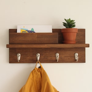 May include: A dark brown wooden wall mounted shelf with a small shelf on top for mail and a lower shelf with four hooks for hanging items. A small potted succulent plant sits on the top shelf. A yellow jacket is hanging on one of the hooks.