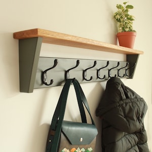 Puede incluir: Un perchero de pared con un estante de madera natural sostenido por dos soportes de color verde oscuro. El panel trasero verde oscuro tiene seis ganchos de metal negro. Un bolso de cuero verde oscuro y una chaqueta acolchada negra cuelgan.