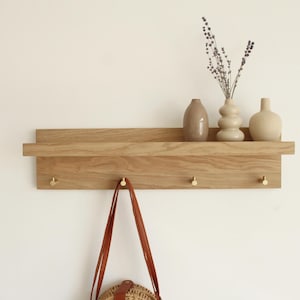 Könnte beinhalten: Wandregal aus Holz mit drei dekorativen Vasen und Trockenblumen. Das Regal hat drei Messing-Haken darunter, an einem hängt eine braune Ledertasche. Ein minimalistisches Design für die Inneneinrichtung.