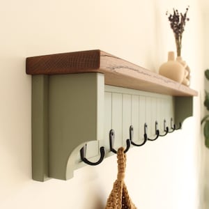 May include: A wall-mounted shelf featuring a natural wood top surface and a sage green base with vertical paneling. The unit includes a row of black metal hooks attached to the underside for hanging items, with a textured tan woven bag hanging from one.