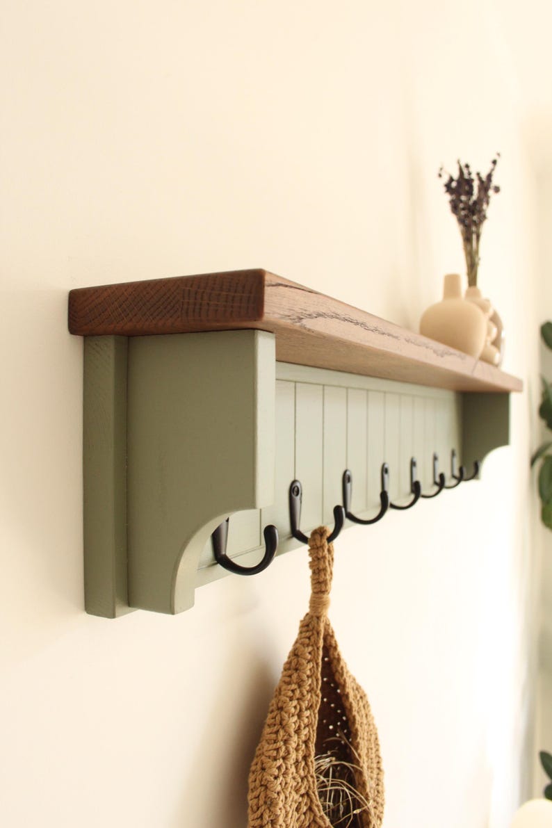 May include: A wall-mounted shelf featuring a natural wood top surface and a sage green base with vertical paneling. The unit includes a row of black metal hooks attached to the underside for hanging items, with a textured tan woven bag hanging from one.