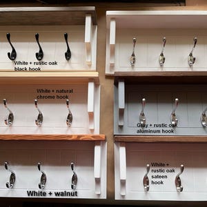 May include: Five different styles of white wooden coat racks with five hooks each. The racks have different wood finishes and hook colors. The finishes are white with rustic oak, white with natural chrome, gray with rustic oak, white with walnut, and white with rustic oak sateen. The hooks are black, chrome, aluminum, and silver.
