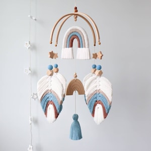 Op de afbeelding: Een houten babymobiel met een regenboog, sterren en veren. De regenboog is gemaakt van garen in tinten blauw, bruin en wit. De veren zijn wit met blauwe en bruine accenten. De sterren zijn van hout en wit geverfd.