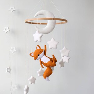 May include: A white felt crescent moon and stars hang from a wooden ring, with a felt orange fox hanging from the bottom. The mobile is suspended from a white ceiling.