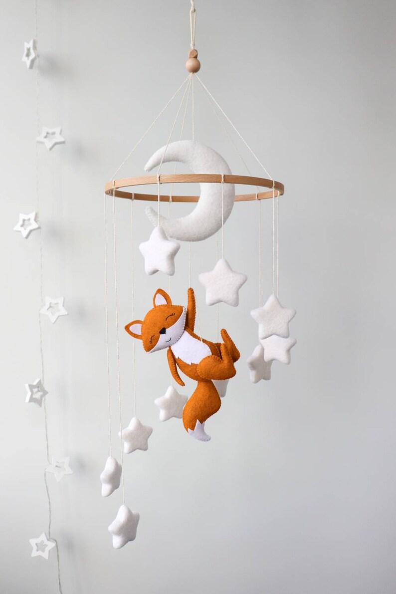 May include: A white felt crescent moon and stars hang from a wooden ring, with a felt orange fox hanging from the bottom. The mobile is suspended from a white ceiling.