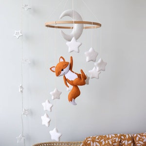 May include: A white felt moon and stars mobile with a felt orange fox hanging from a wooden ring. The mobile is hanging above a wicker bassinet with a mustard yellow and white patterned blanket.