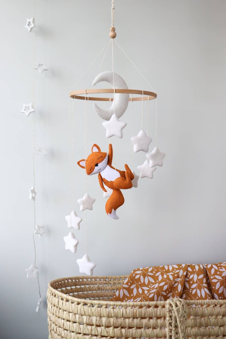 May include: A white felt moon and stars mobile with a felt orange fox hanging from a wooden ring. The mobile is hanging above a wicker bassinet with a mustard yellow and white patterned blanket.