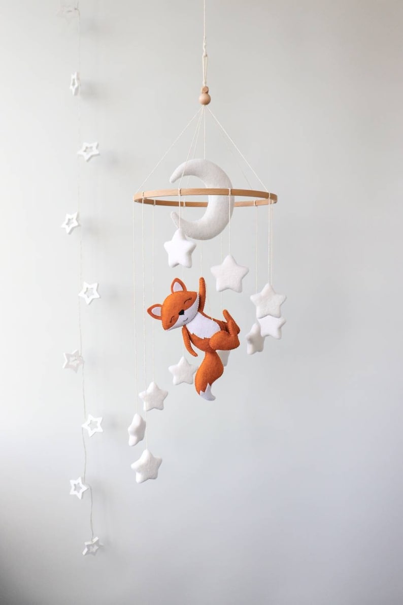 May include: A wooden baby mobile with a white felt crescent moon and white felt stars. A felt orange fox hangs from the mobile, holding onto a white felt star.
