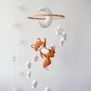 May include: A white felt moon and stars mobile with a brown felt fox hanging from a wooden ring. The mobile is suspended from the ceiling.