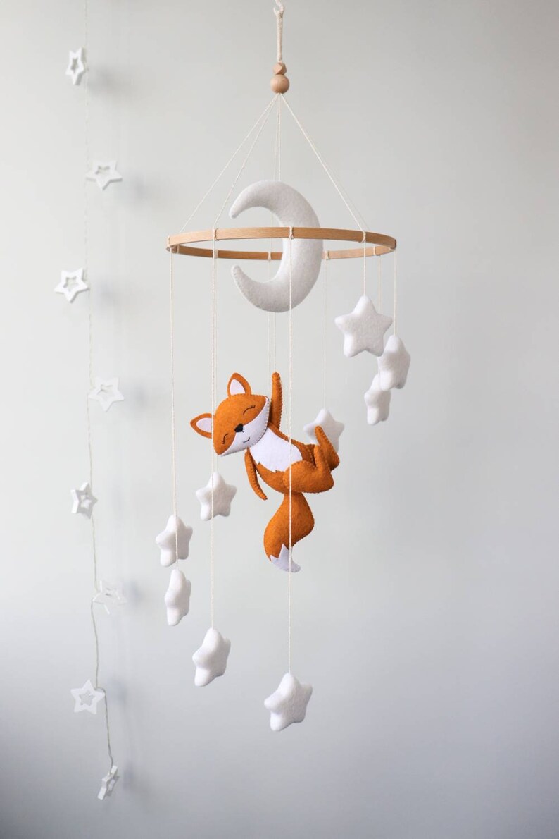 May include: A white felt moon and stars mobile with a brown felt fox hanging from a wooden ring. The mobile is suspended from the ceiling.