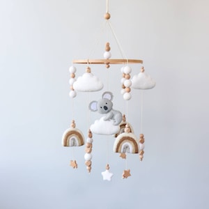 May include: A wooden baby mobile with a gray koala bear, white clouds, rainbow-colored felt rainbows, and wooden beads. The mobile is hanging from a white ceiling.
