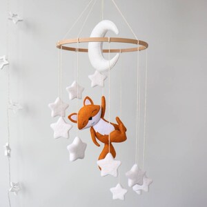 May include: A white felt moon and stars hang from a wooden ring, with a brown felt fox hanging from the bottom. The mobile is suspended from a white ceiling.