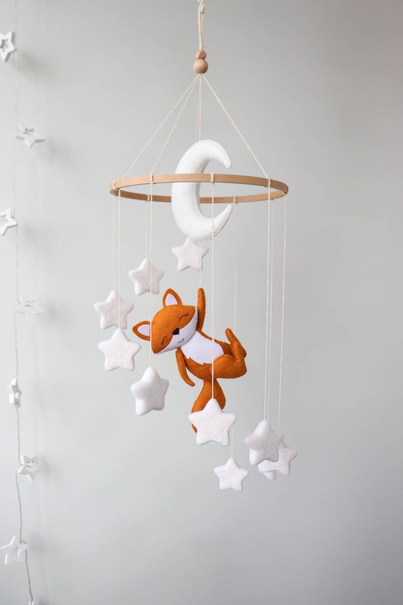 May include: A white felt moon and stars hang from a wooden ring, with a brown felt fox hanging from the bottom. The mobile is suspended from a white ceiling.