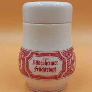 Może przedstawiać: Biały ceramiczny słoik z pokrywką. Słoik ma czerwony ozdobny pasek ze słowami "Rüdesheimer Früchtetopf" w czerwonym skrypcie. Słoik jest na pomarańczowym tle.